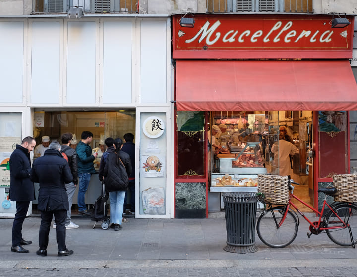 Foto di "La Ravioleria Sarpi" di Milano con una fila di clienti davanti, e la macelleria alla sua destra.